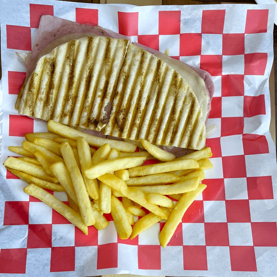 Grilled panini sandwich with ham and cheese, served with golden french fries on checkered paper.