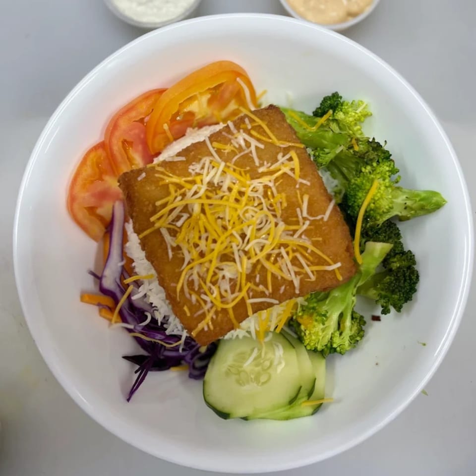A white bowl filled with a breaded chicken cutlet topped with cheese, broccoli, tomatoes, and cucumber.