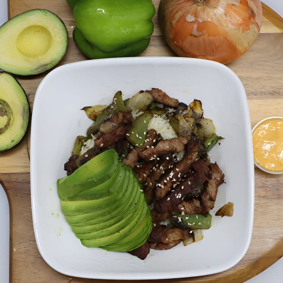 A white bowl of steak, peppers, onions, rice, and avocado, with sauce on a wooden board.