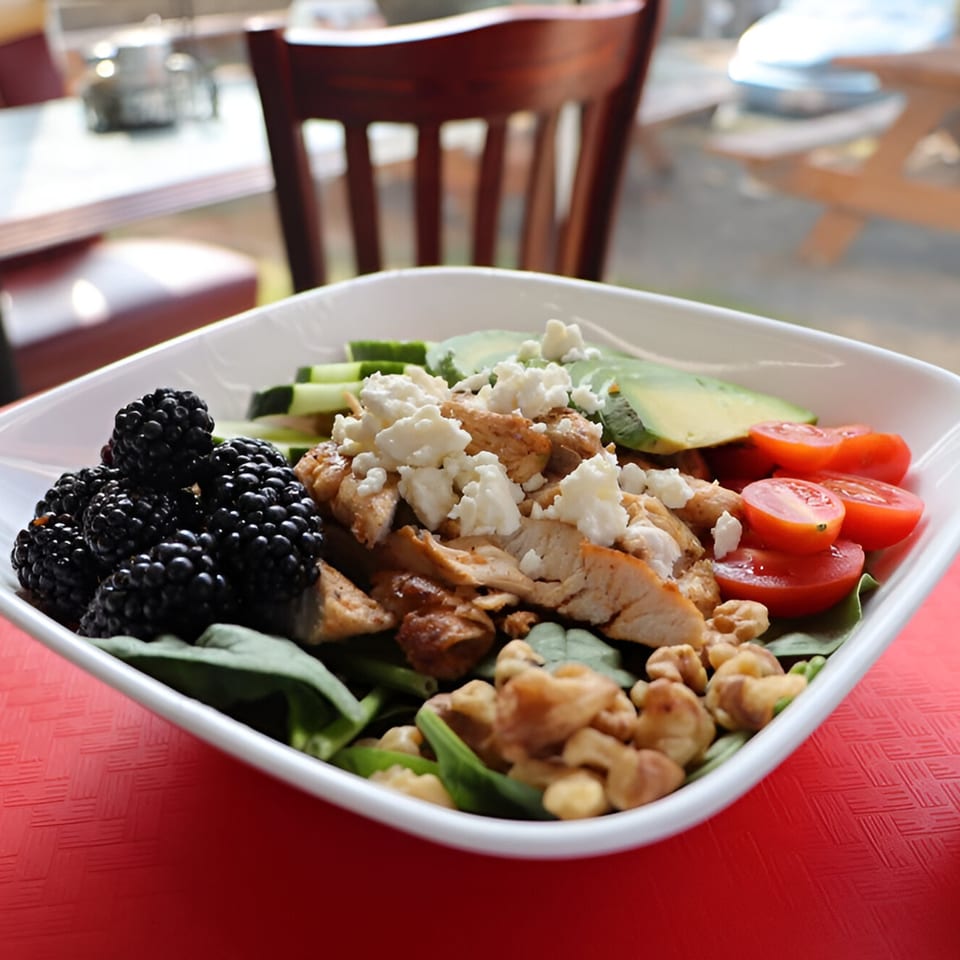 A white bowl of salad with chicken, blackberries, avocado, tomatoes, feta, and walnuts.
